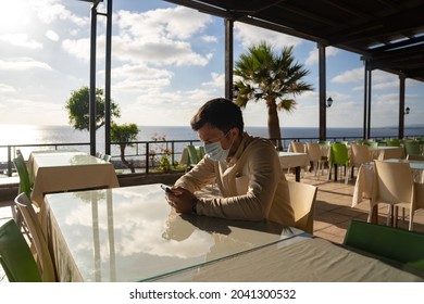 Lonely Sad Masked Man Sits And Uses Smartphone Himself In Empty Restaurant In Cyprus At Agios Georgios. Visitor Wearing Mask Waits Order While Sitting In Deserted Cafe, Social Distance And Quarantine.