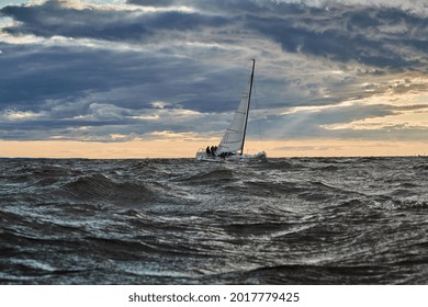 The lonely sailboat on the horizon in sea at sunset, the storm sky of different colors, big waves, sail regatta, cloudy weather, only main sail, sun beams
