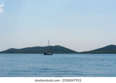 Lonely sailboat resting on still blue water with green hills in the distance under cloudless sky on warm sunny day. Concept of relaxation, maritime exploration, and scenic beauty. High quality photo - Powered by Shutterstock