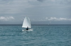 Lonely Sailor On Training Sailing Pram Optimist Education Boat In The Sea, Water Background And Cloudy Sky