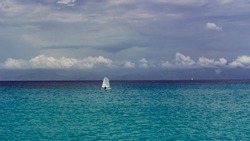 Lonely Sailor On Training Sailing Pram Optimist Education Boat In The Sea, Water Background And Cloudy Sky