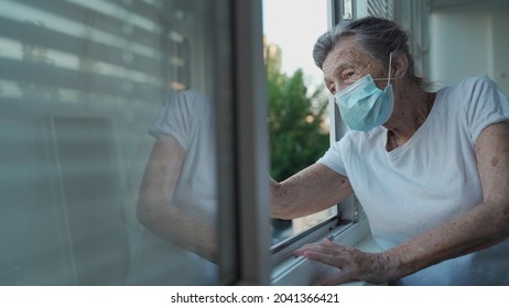 Lonely Senior Woman In Mask Stands By Window And Waves Her Hand To Family Who Came Visit Her At Hospital. 90 Year Old Female In Disguise Says Goodbye To Her Relatives In Nursing Home.