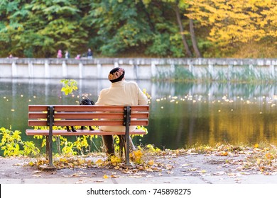 Lonely senior woman with a small black dog sitting on a bench by the autumn lake or river in a city park. Relaxation. Loneliness concept. 