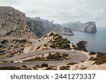 Lonely small white car driving by serpentine curved asphalt mountain road near Lighthouse of Cap de Formentor with beautiful seascape with rocky coast. Mallorca Island, Balearic Islands, Spain.