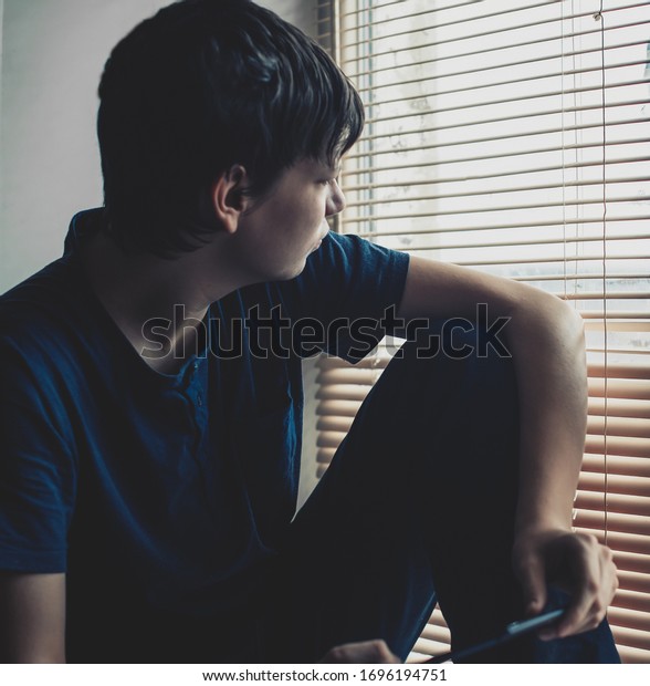 A lonely, squeezed teenager with social problems\
sits sadly on the windowsill in an apartment and looks out the\
window with blinds.
