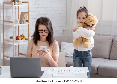 Lonely teen girl hugging teddy bear while her busy mom working online from home, not paying attention to child. Depressed kid feeling neglected, missing her parent. Covid-19 quarantine family problems