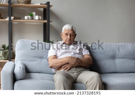 Lonely thoughtful older 75s man sits alone on sofa thinking of loneliness, life or health problems. Melancholic senior lost in sad thoughts looks pensive, recollect past, suffers from solitude concept