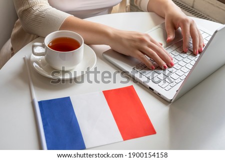 Lonely woman freelancer with flag of France, enjoying having breakfast with cup of coffee working on laptop sitting near window in cafe at morning. 