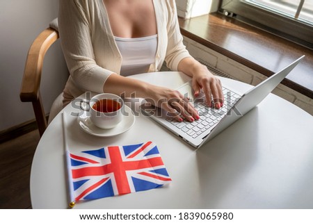 Lonely woman freelancer with flag of Great Britain enjoying having breakfast with cup of coffee working on laptop sitting near window in cafe at morning. 