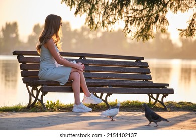 Lonely woman sitting on lake side bench enjoying warm summer evening. Solitude and relaxation concept.