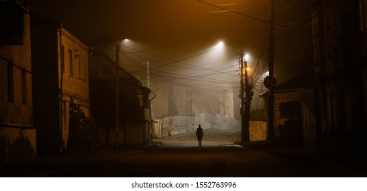 Lonely Woman Walking In Foggy Old City With Street Lights In A Coat