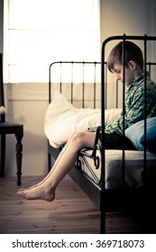 Lonely Young Boy Sitting on his Bed Inside his Room with Hanging Feet, Showing Pensive Facial Expression.