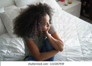 Lonely young latina woman sitting on bed. Depressed hispanic girl at home, looking away with sad expression.