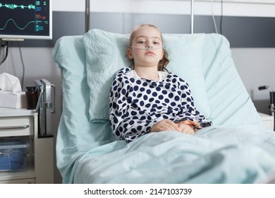 Lonely Young Pale Sick Girl Patient Resting In Pediatric Clinic Bed While In Recovery Ward Room. Ill Child Sitting Alone In Hospital Bed While Wearing Nasal Oxygen Tube For Life Support.