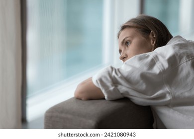 Lonely young woman lies on sofa, looking out window, having melancholic mood, suffer of unrequited love, break up, feel depressed, unhappy alone at home, pensive attractive female thinks about problem