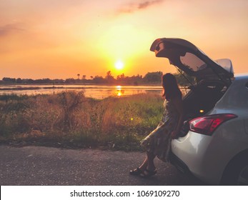 Lonely Young Woman Traveler Sitting On Hatchback Car And Looking At The Sunset Against Orange Sky Background And Sunlight Reflection On Lake. Travel In Holiday Concept