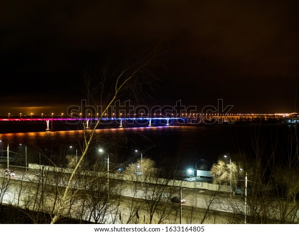 long automobile bridge\
over large river illuminated by multi-colored lights. Cars driving\
along embankment of river. little traffic late in evening. Trees in\
foreground