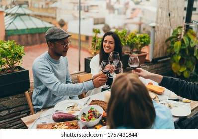 Long Awaited Meeting Of Best Friends On Summer Rooftop With Cozy Garden, Multicultural Group Of People Making Cheers With Red Wine Glasses Laughing And Joking While Celebrating Meeting Reunion Outside