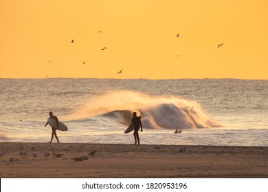 Long Beach, New York - September 21, 2020 : Silhouette of a surfers walking on the beach at sunset, with waves crashing in the background.