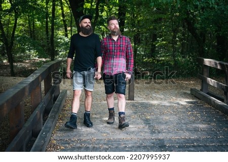 Long bear hipster gay couple walking in park toward camera, crossing wooden bridge and holding hands. Good looking homosexuals outdoor in autumn. 