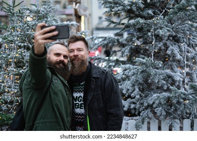 Long beard hipsters homosexuals enjoying Christmas advent, smiling and taking selfie in front of Xmas tree outdoor. Attractive male gay couple celebrating holiday season.  - Powered by Shutterstock