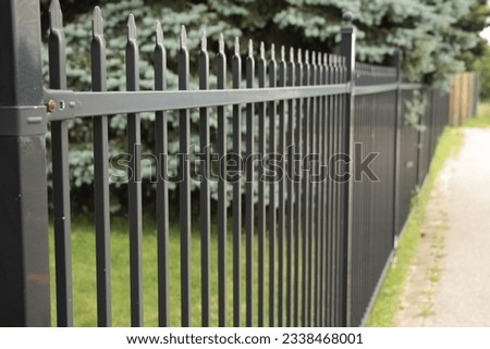 long black metal bar fence with pine tree and grass behind it, top of fence in shot with wooden fence in background