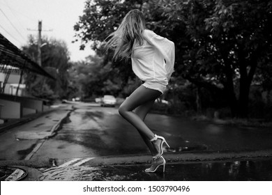 Long Blonde Hair Shaking And Wet After The Summer Pouring Rain. Lady Having Fun Outdoor On The Street In Her Man's Shirt And On High Heel Shoes With Open Toes. Puddles And Road With Water Streams.