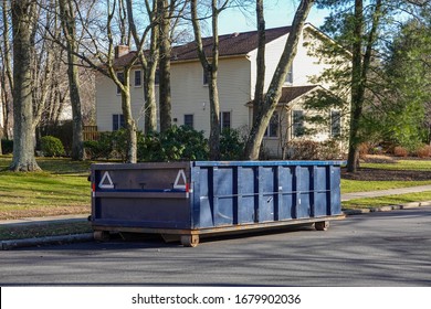 Long blue dumpster on a street by a curb behind a residential home