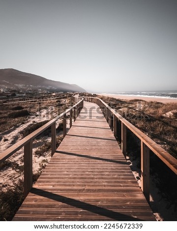 A long boardwalk on a grassy sand dune at the beach leading to the mountain and ocean with desaturated colors. The way ahead concept, Quiaios Beach, Portugal