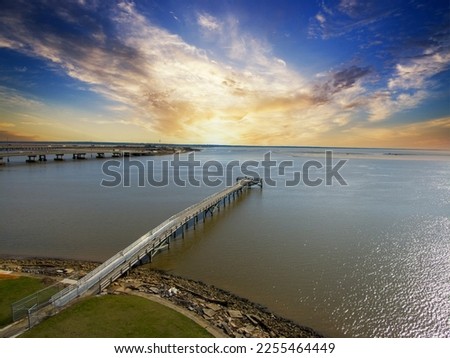 A long brown wooden pier of the rippling waters of Mobile Bay with cars driving on the highway and powerful clouds at sunset at USS Alabama Battleship Memorial Park in Mobile Alabama USA