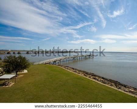 A long brown wooden pier of the rippling waters of Mobile Bay with cars driving on the highway and a gorgeous blue sky with clouds at USS Alabama Battleship Memorial Park in Mobile Alabama USA