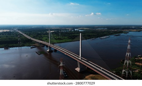 Long cable-stayed bridge spans wide river under clear blue sky. Tall towers support the bridge's cables. Boats navigate calm river waters. Green fields stretch to distant horizon