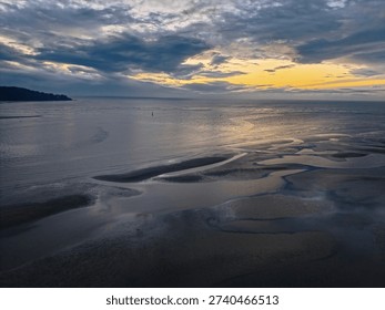 🌊 Long Description
The image presents a dramatic black-and-white seascape captured at what appears to be dusk or early evening. The sky dominates the upper half of the composition, filled with thick, layered clouds stretching across the horizon. The clouds appear heavy and textured, creating a sense of depth and movement. Light breaks subtly through the cloud cover near the horizon, casting a soft, diffused glow across the water below.

The sea is calm and expansive, extending toward the distant horizon where it meets the sky in a seamless gradient of grey tones. Gentle ripples shimmer faintly, reflecting the muted light from above. On the left side of the frame, a dark silhouette of a landmass or small hill juts into the water, providing contrast and anchoring the composition.

In the foreground, the shoreline reveals intricate tidal patterns in the sand. Shallow pools of water have formed smooth, organic shapes that curve and flow naturally, creating abstract patterns against the darker wet sand. These reflective pools mirror parts of the sky, adding texture and visual interest. The contrast between light and shadow emphasizes the smooth surfaces of the water and the subtle contours of the sand.

The monochrome treatment enhances the mood, giving the scene a timeless and contemplative atmosphere. Without color distractions, attention is drawn to texture, contrast, light, and form. The overall feeling is peaceful yet slightly dramatic—evoking solitude, reflection, and the quiet power of nature at the edge of day.
