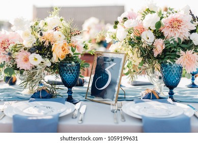 Long dinner tables covered with white cloth, served with porcelain and blue glasses and rich decorated with flowers stand on the roof of a house