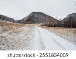 A long dirt road that winds its way through a picturesque snowy field, with majestic mountains soaring in the background, creating a stunning natural landscape that captivates the eyes