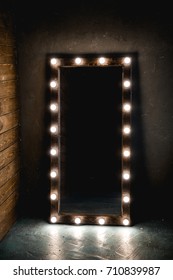 Long dressing mirror with light bulbs stands on the floor against a wooden and concrete wall