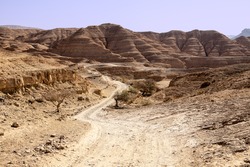 A Long Dusty Road Winds Through The Isolated And Remote Hills And Valleys Of The Negev Desert. In The Background, Hills Of Sedimentary Sandstone Loom Over The Road.