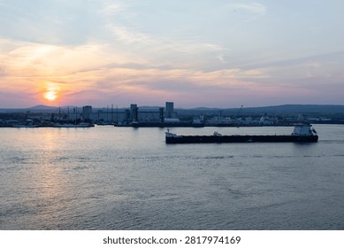 Long empty container ship on the St. Lawrence river seen during a pink summer sunset, with large sailboats moored in the port and the old town in silhouette in the background