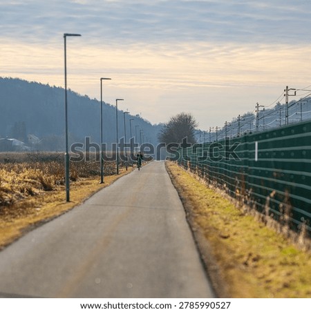 Long empty cycling path with distant rider beside railway fence under calm cloudy sky