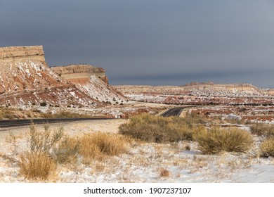 Long empty highway cutting through red rock valley covered in snow with mountains and overcast sky in rural New Mexico