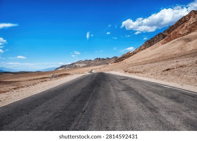 A long, empty highway stretching through the desert landscape of Death Valley, California, with distant mountains and vast blue sky, symbolizing travel, isolation, and the open road in the American West.