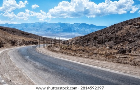 A long, empty highway stretching through the desert landscape of Death Valley, California, with distant mountains and vast blue sky, symbolizing travel, isolation, and the open road in the American West.