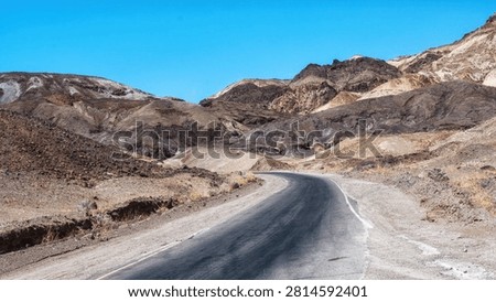 A long, empty highway stretching through the desert landscape of Death Valley, California, with distant mountains and vast blue sky, symbolizing travel, isolation, and the open road in the American West.