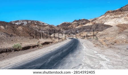 A long, empty highway stretching through the desert landscape of Death Valley, California, with distant mountains and vast blue sky, symbolizing travel, isolation, and the open road in the American West.