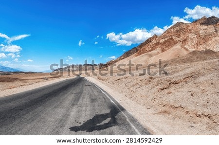 A long, empty highway stretching through the desert landscape of Death Valley, California, with distant mountains and vast blue sky, symbolizing travel, isolation, and the open road in the American West.