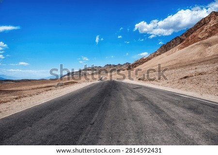 A long, empty highway stretching through the desert landscape of Death Valley, California, with distant mountains and vast blue sky, symbolizing travel, isolation, and the open road in the American West.