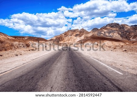 A long, empty highway stretching through the desert landscape of Death Valley, California, with distant mountains and vast blue sky, symbolizing travel, isolation, and the open road in the American West.