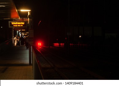A long empty railway with a foreboding red light at the end. Waiting passengers wait for the next incoming train.