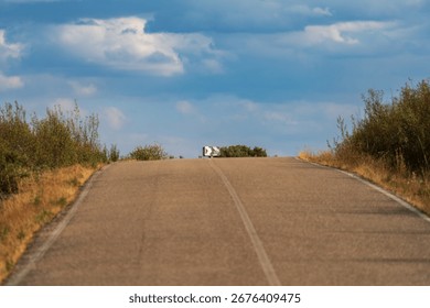 A long, empty road extends toward the horizon beneath a wide, cloudy sky, bordered by vegetation on both sides.