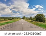 A long, empty road with a few trees in the background. The sky is cloudy, but the sun is still shining through